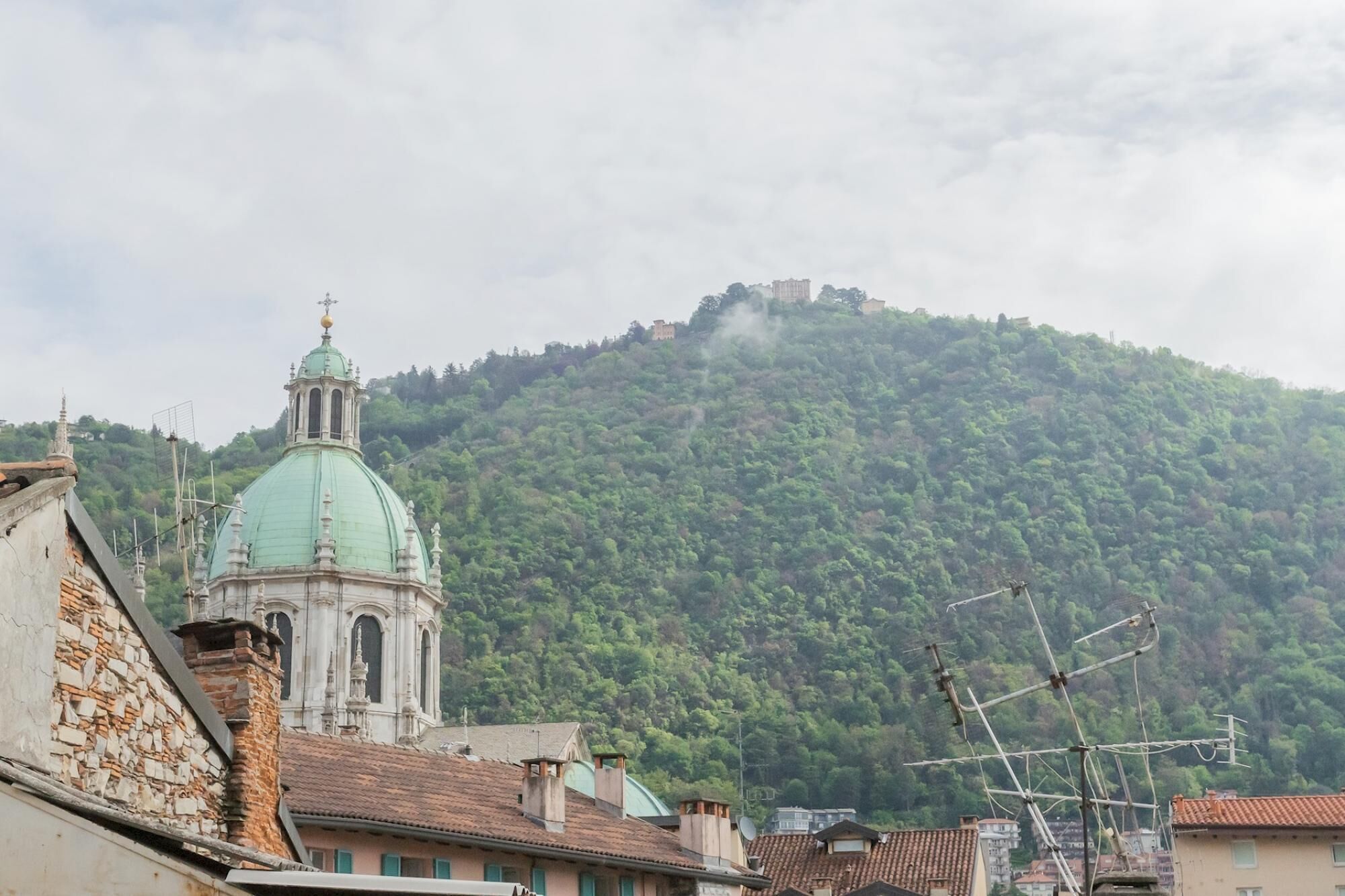 A building with a green dome and a bell tower, surrounded by residential buildings and a hill with a castle-like structure.