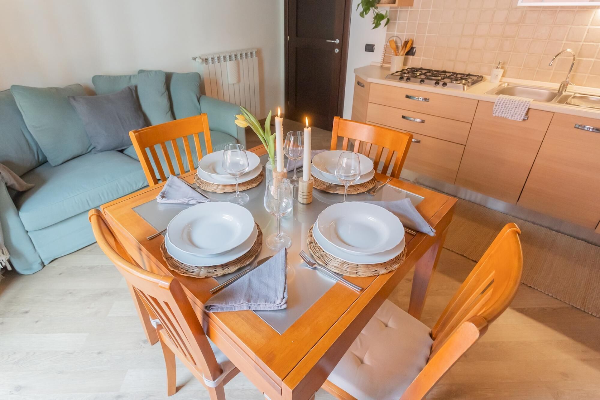 Taula de menjador posta amb plats, gots i espelmes, envoltada de caires de fusta en una habitació amb un sofà i una cuina al fons.