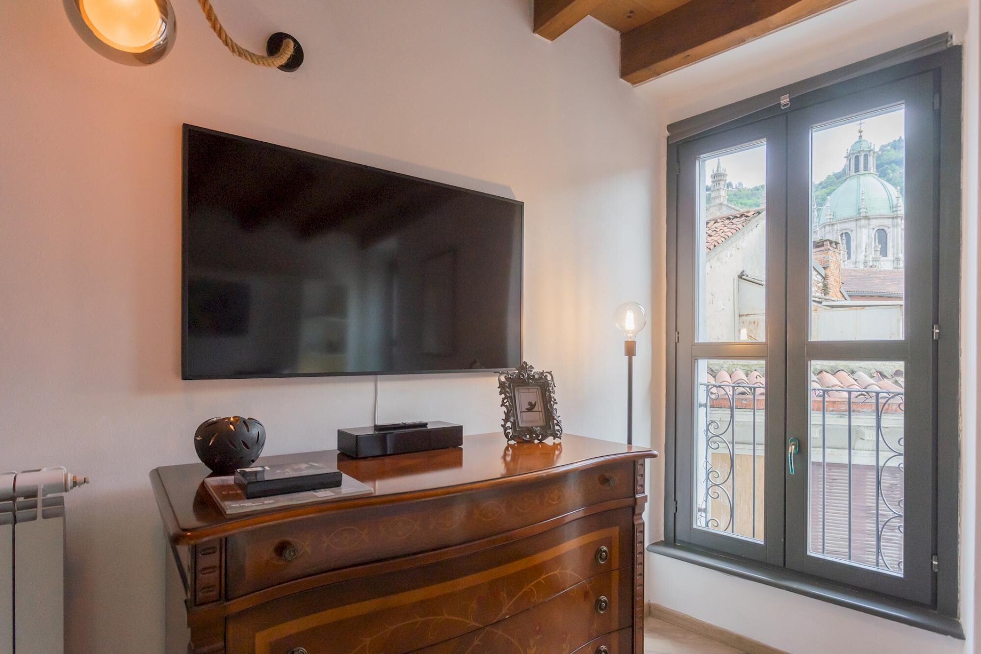 Une pièce dotée d’un grand téléviseur à écran plat fixé au mur, d’une commode en bois avec des tiroirs, d’une petite table ornée d’un cadre décoratif et d’une fenêtre donnant sur un immeuble et un balcon.