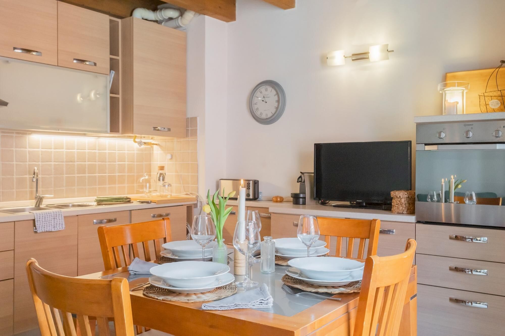 A modern kitchen with wooden cabinets, a dining table set for four, a clock, and a television.