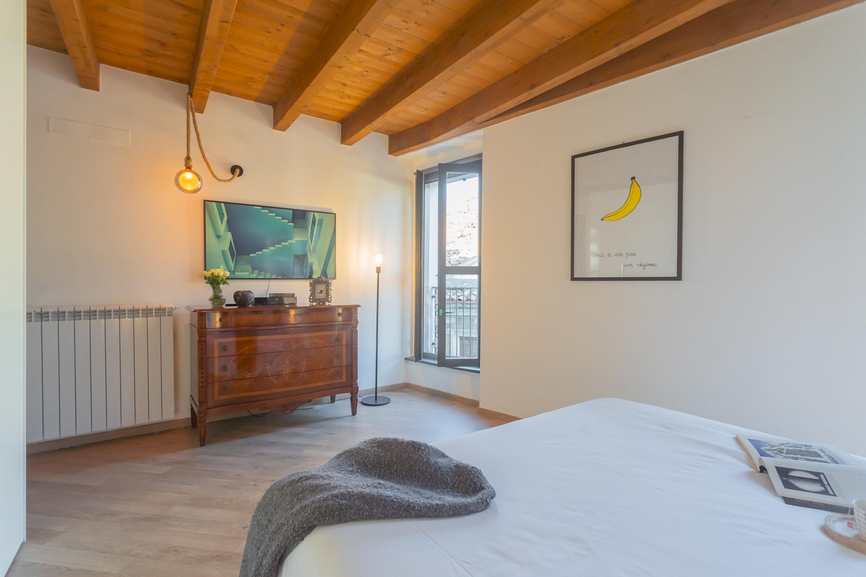 A bedroom with a wooden ceiling, a bed with a white sheet, a framed picture of a banana, a dresser, and a painting on the wall.