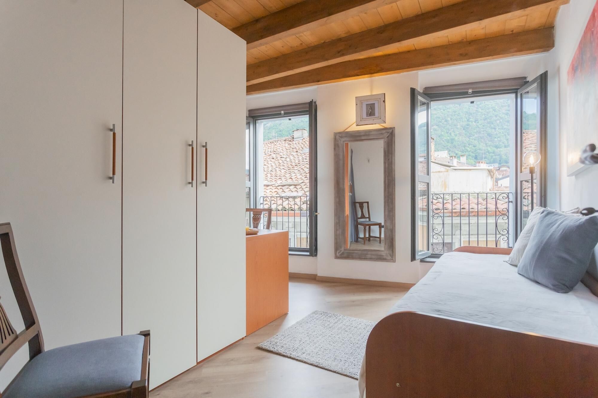 A bedroom with a bed, a chair, a wardrobe, a desk, and a balcony with a view of the city.