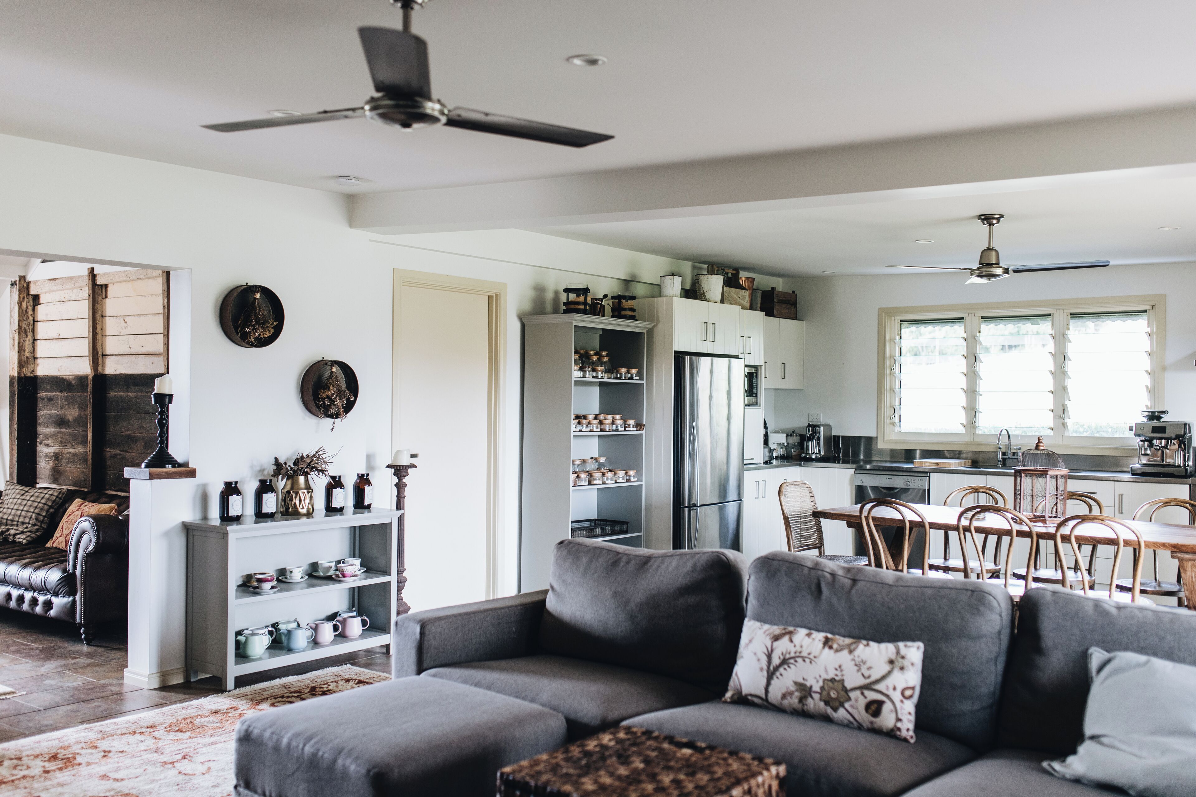 A modern living room with a grey sofa, a dining area with wooden chairs, a kitchen with white cabinets, and a ceiling fan.