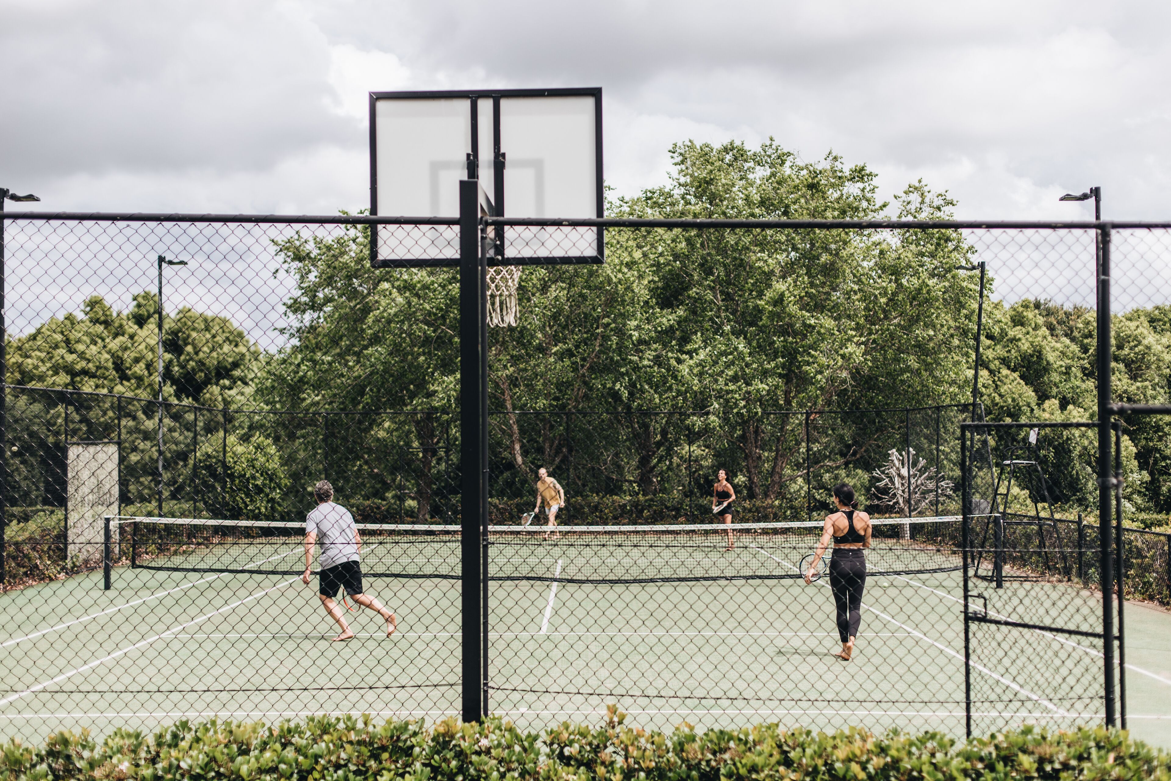 A tennis court with a chain-link fence, a basketball hoop, and trees in the background.