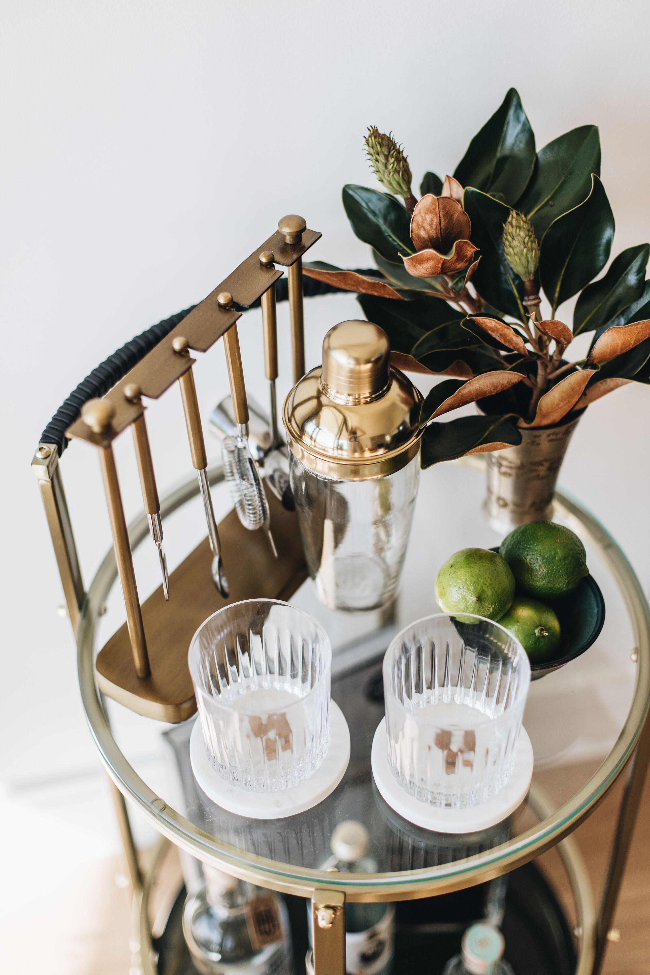 A round glass table with a gold metal frame, holding a cocktail shaker, two clear glasses, and a small vase with green leaves and a brown object.