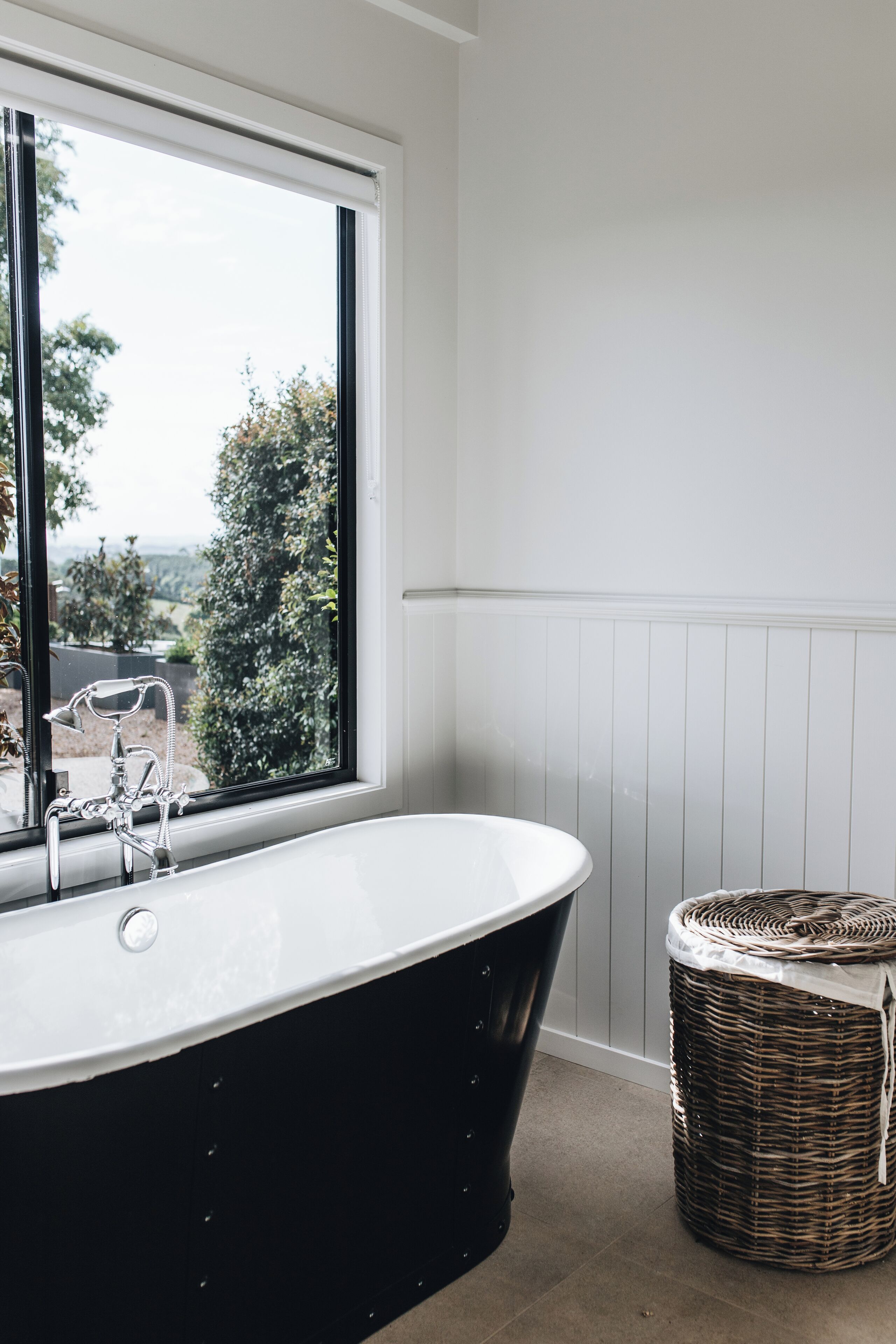 A bathroom with a freestanding bathtub, a wicker basket, and a window with a view of trees and a car.
