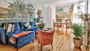 A living room with a blue sofa, a pink armchair, a dining table with chairs, and a large painting of a tropical landscape. - Appartement Chernoviz - Welkeys (Paris)
