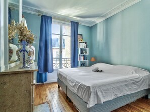 Ein Schlafzimmer mit Bett, Kommode, Fenster mit Vorhängen, Bücherregal und Lampe.