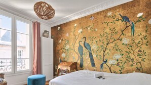 A bedroom with a large bed, a wooden floor, a window with curtains, and a wall mural of birds and flowers. - Appartement Chernoviz - Welkeys (Paris)