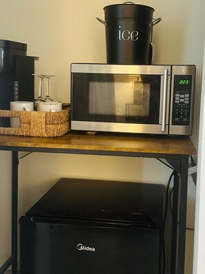 Un horno microondas con pantalla digital, una olla negra etiquetada como “hielo” encima y una bandeja de mimbre con dos tazas blancas.