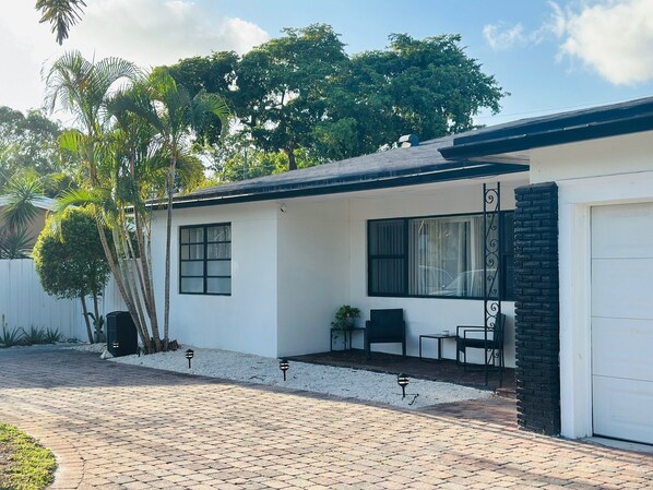Una casa de un piso con fachada blanca, techo negro y entrada de acceso pavimentada. Cuenta con un pequeño patio con mesa y sillas, y un portón de garaje a la derecha.