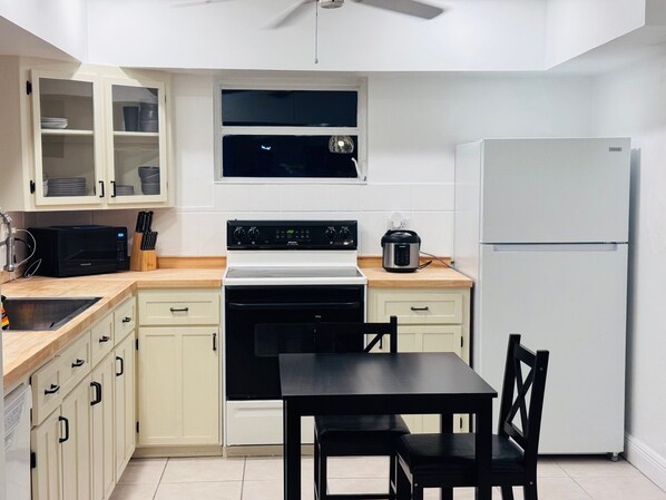 Una cocina compacta con armarios blancos, estufa negra, microondas, refrigerador y una mesa de comedor con sillas.