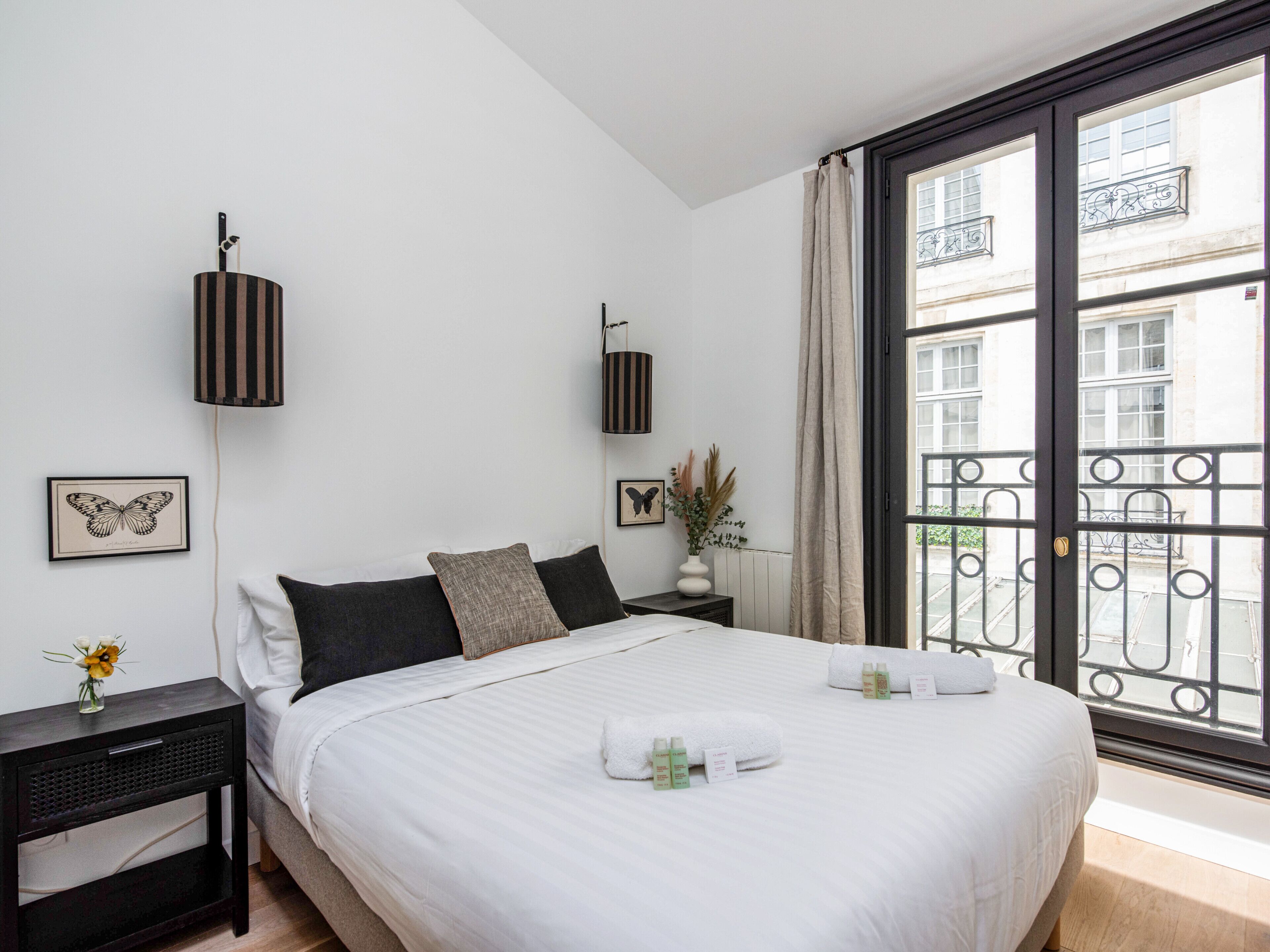 Une chambre d’hôtel dotée d’un grand lit, de deux tables de chevet, d’une fenêtre donnant sur la ville et d’un balcon agrémenté d’une rambarde en fer forgé.