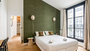 Quarto moderno com uma cama grande, uma parede verde, duas luminárias de cabeceira, vista para a varanda externa e piso de madeira.