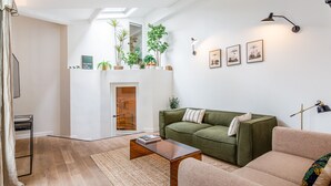 Uma sala de estar moderna com um sofá verde, uma poltrona bege e uma mesa de centro de madeira. Há várias plantas em uma estante, um quadro na parede e um claraboia sobre a lareira.