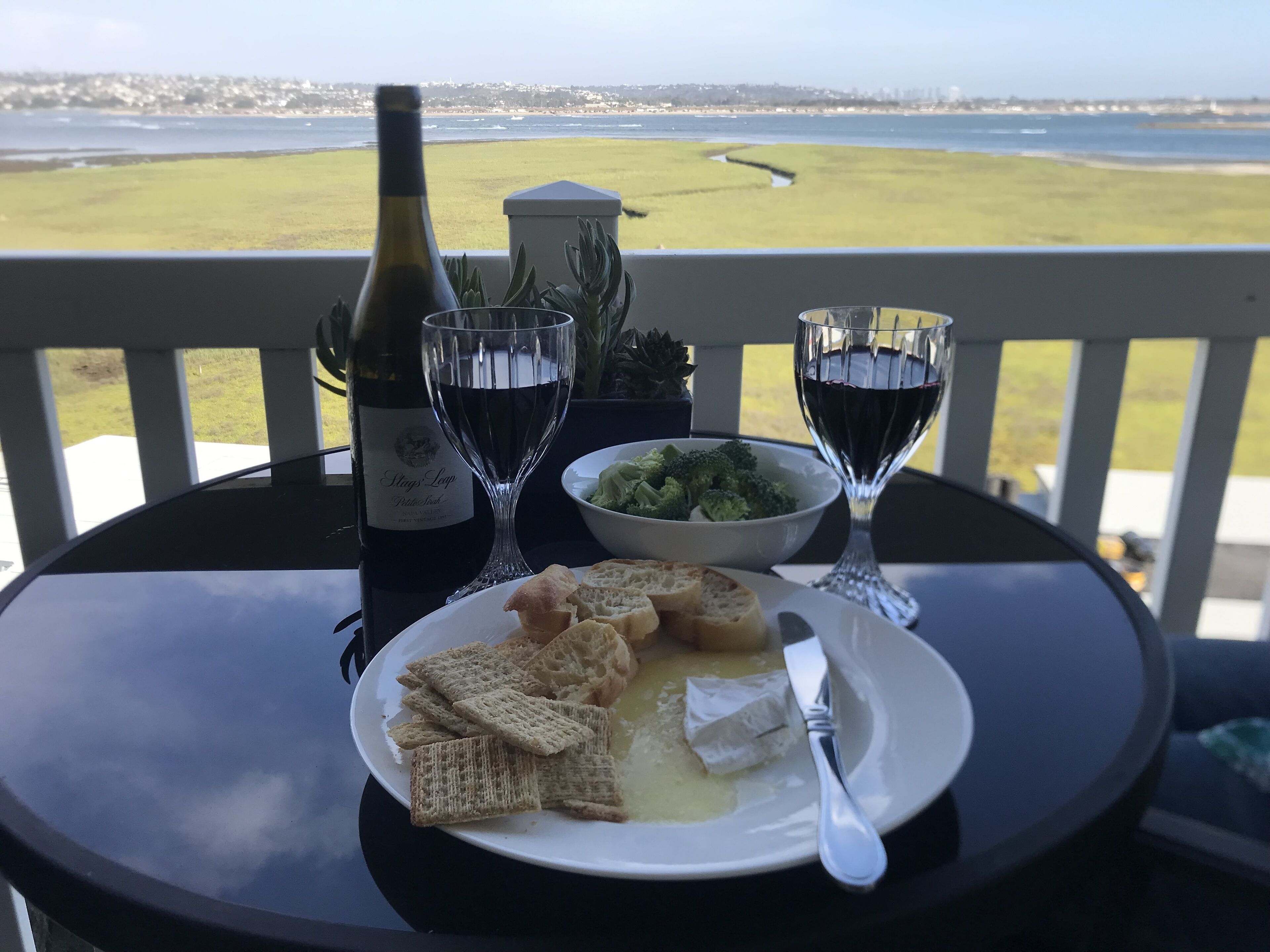 A table with a bottle of wine, two wine glasses, a bowl of broccoli, a plate with crackers and cheese, and a knife on a balcony overlooking a beach.