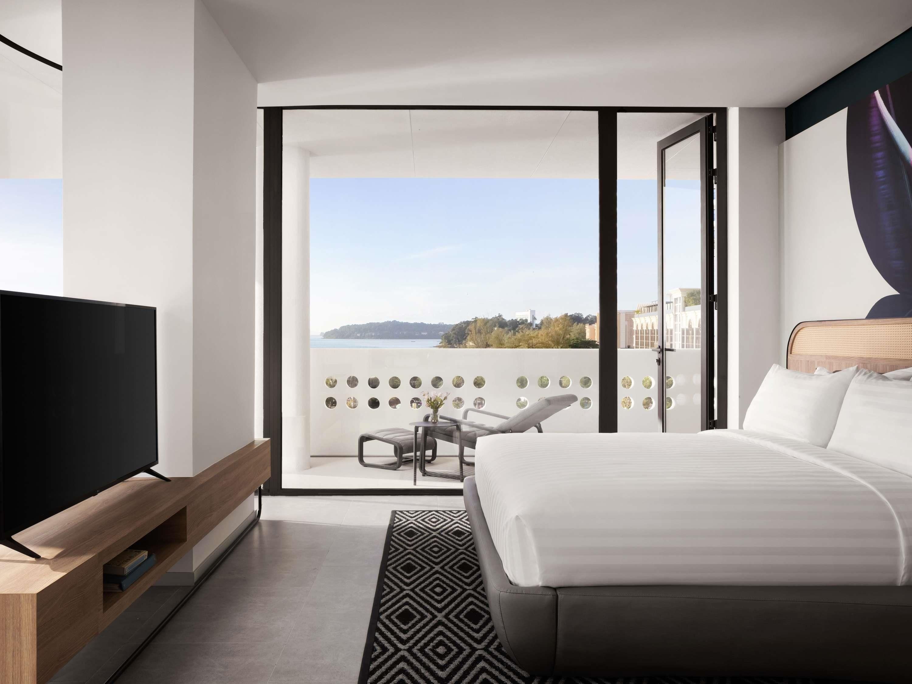 A modern hotel room with a large bed, a TV on a wooden stand, a balcony with a view, and a patterned rug.