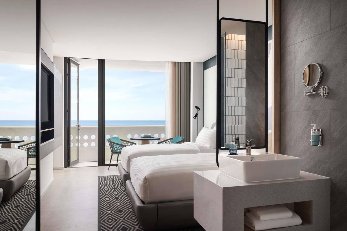 A modern hotel room with a large bed, a bathroom with a sink and mirror, a balcony with a view of the sea, and a patterned rug.