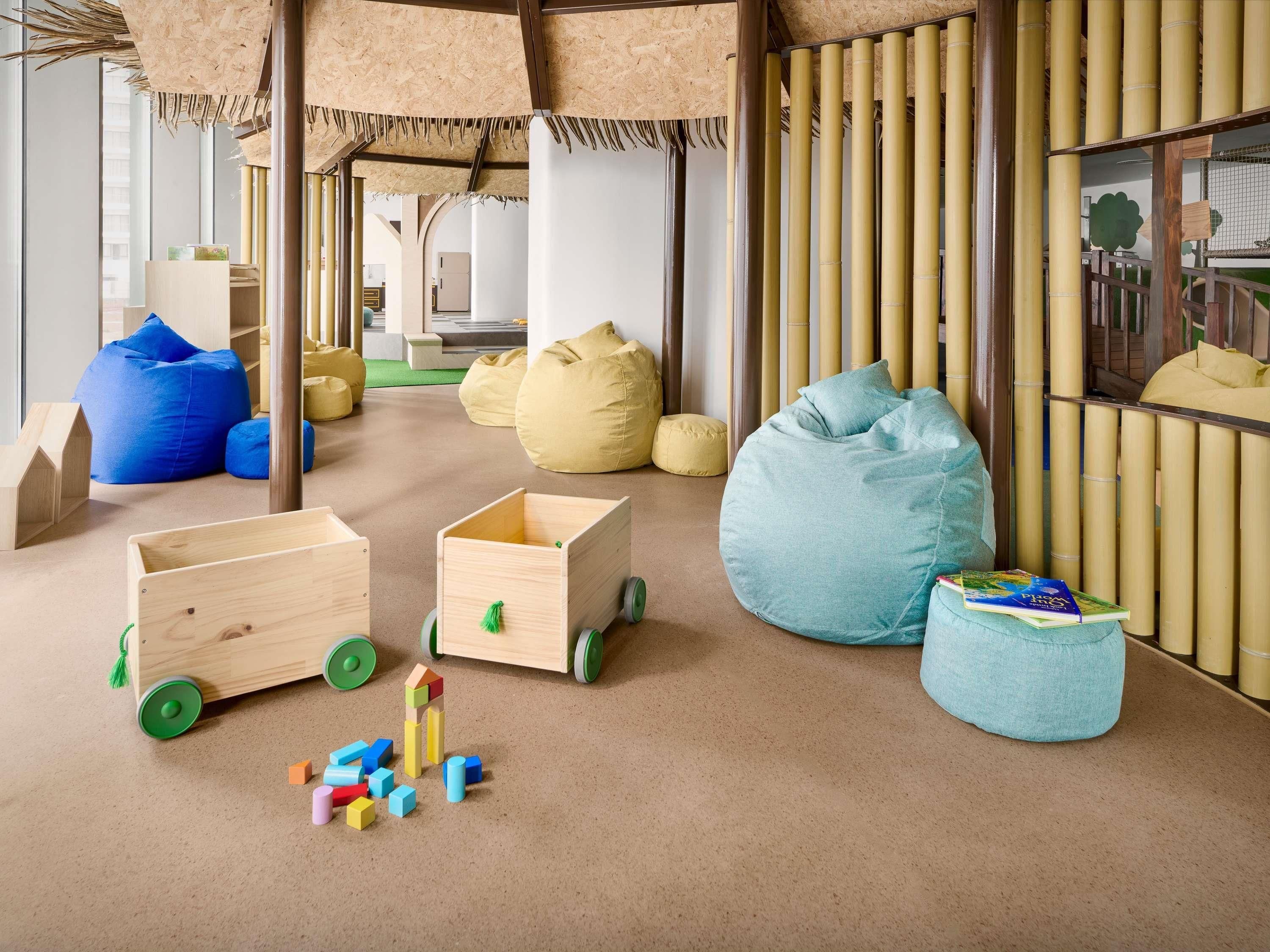 Uma sala de brinquedos com casinhas de madeira, puffs e uma estante de livros.