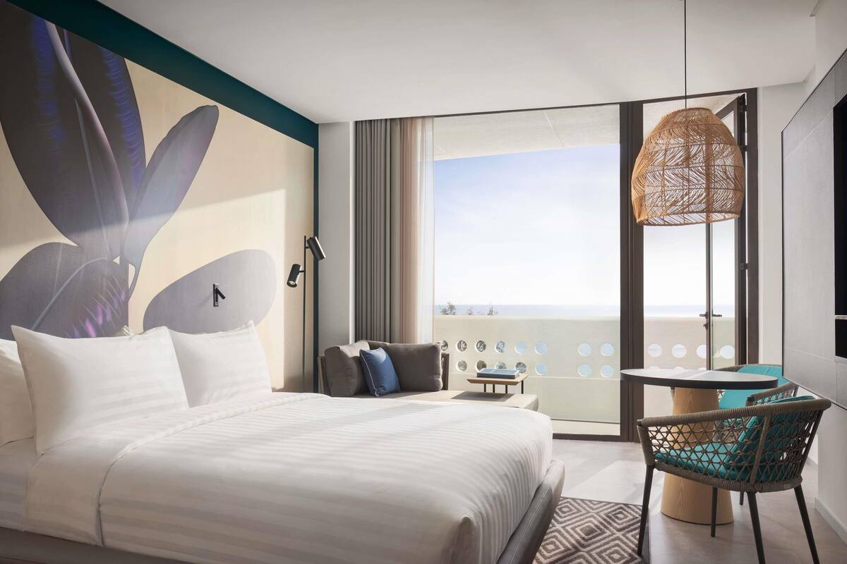 A hotel room with a bed, a chair, a table, a large window with a view, and a decorative wall piece.