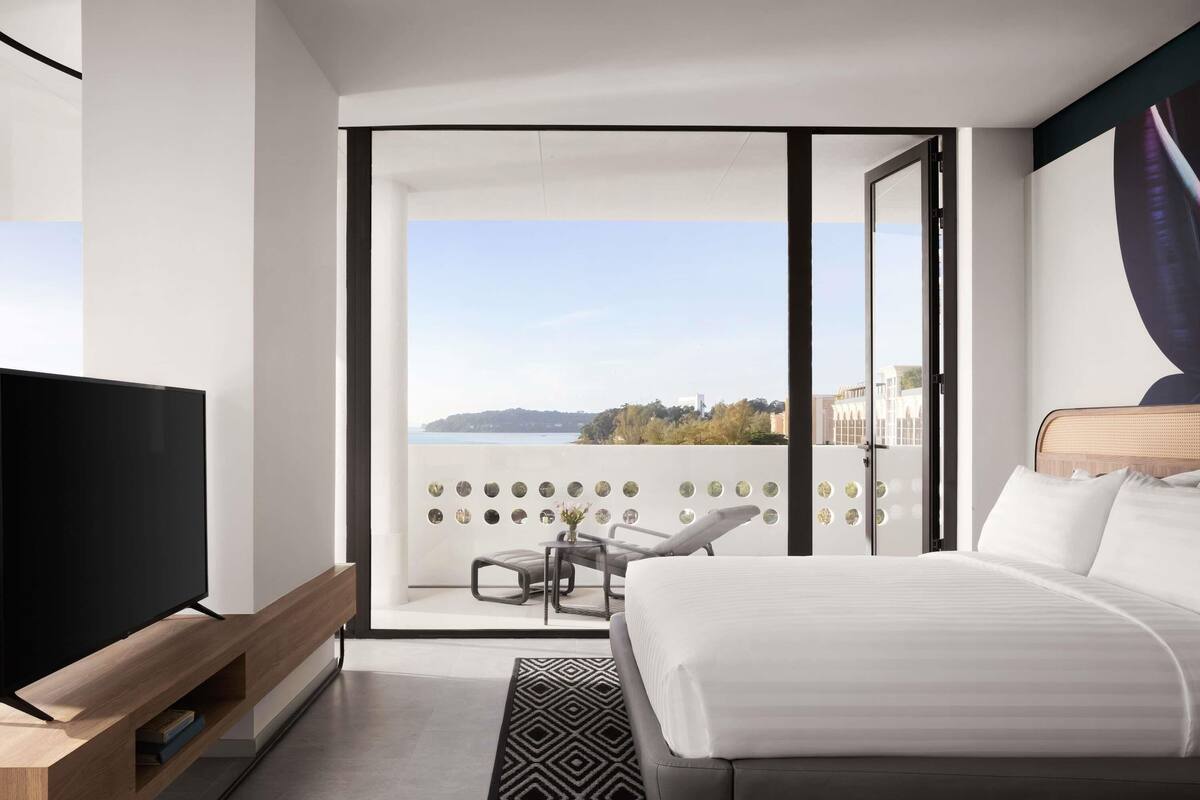A modern hotel room with a large bed, a TV on a wooden stand, a balcony with a view, and a patterned rug.