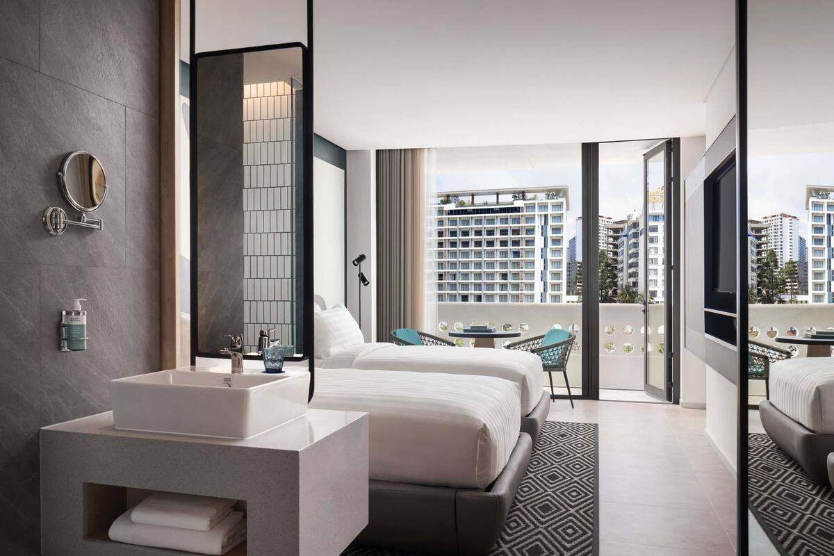 A modern hotel room with a large bed, a separate bathroom with a vanity, a balcony with a view of the city, and a large mirror.