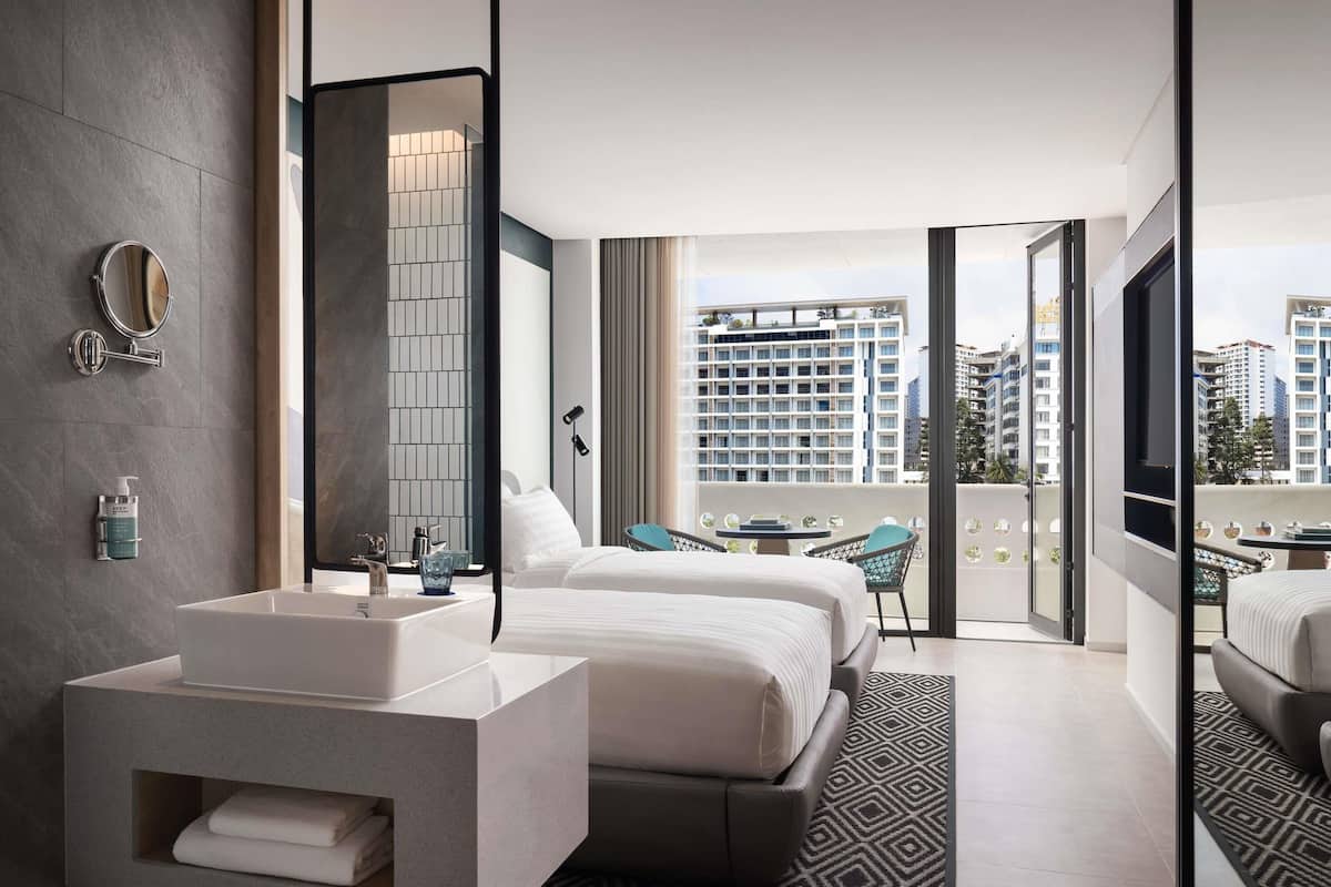 Ett modernt hotellrum med en stor säng, ett separat badrum med handfat, en balkong med utsikt över staden och en stor spegel.