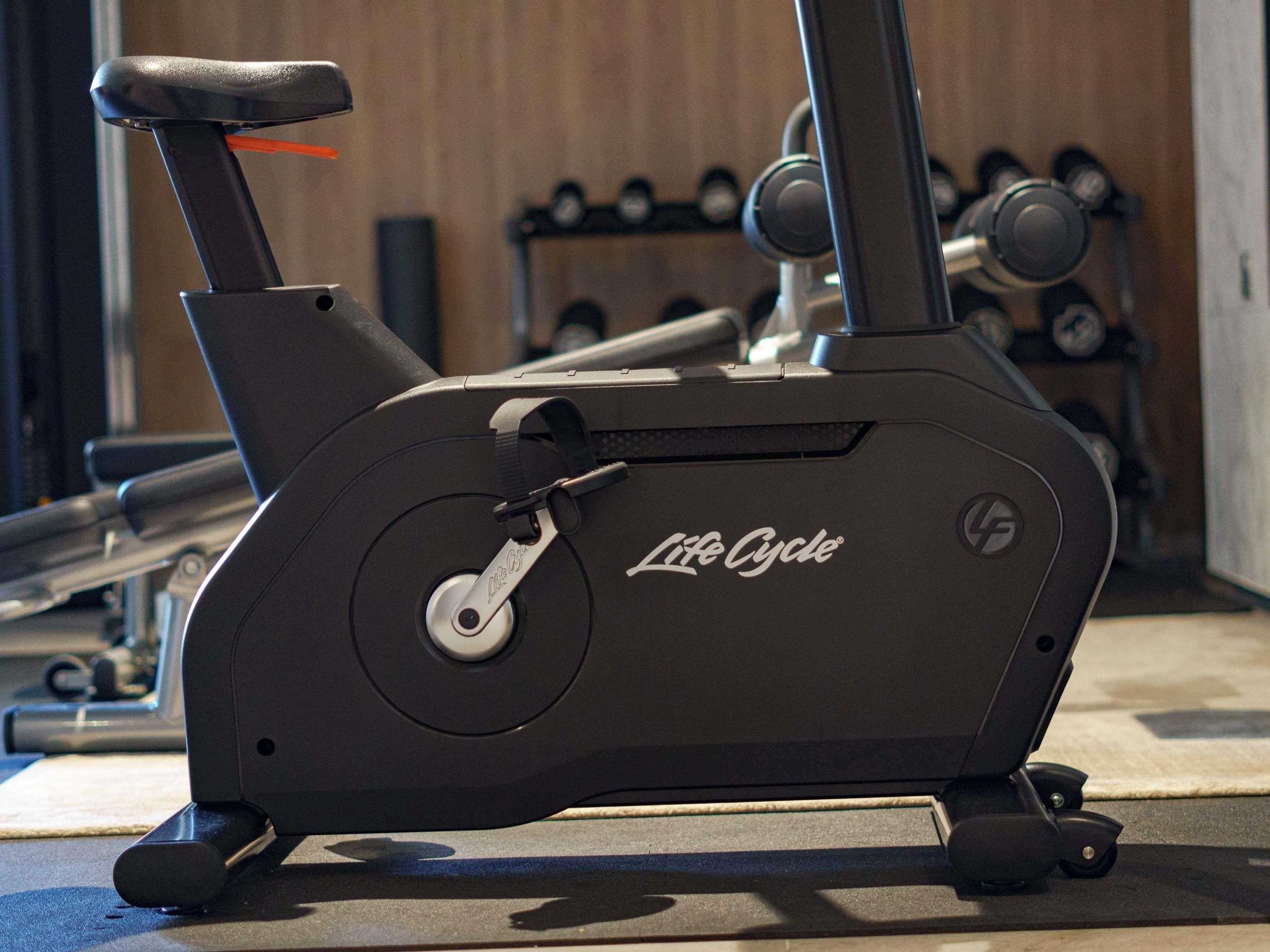 En stillastående træningscykel med Life Cycle logo, placeret i et fitnesscenter med en trævæg og håndvægte i baggrunden.
