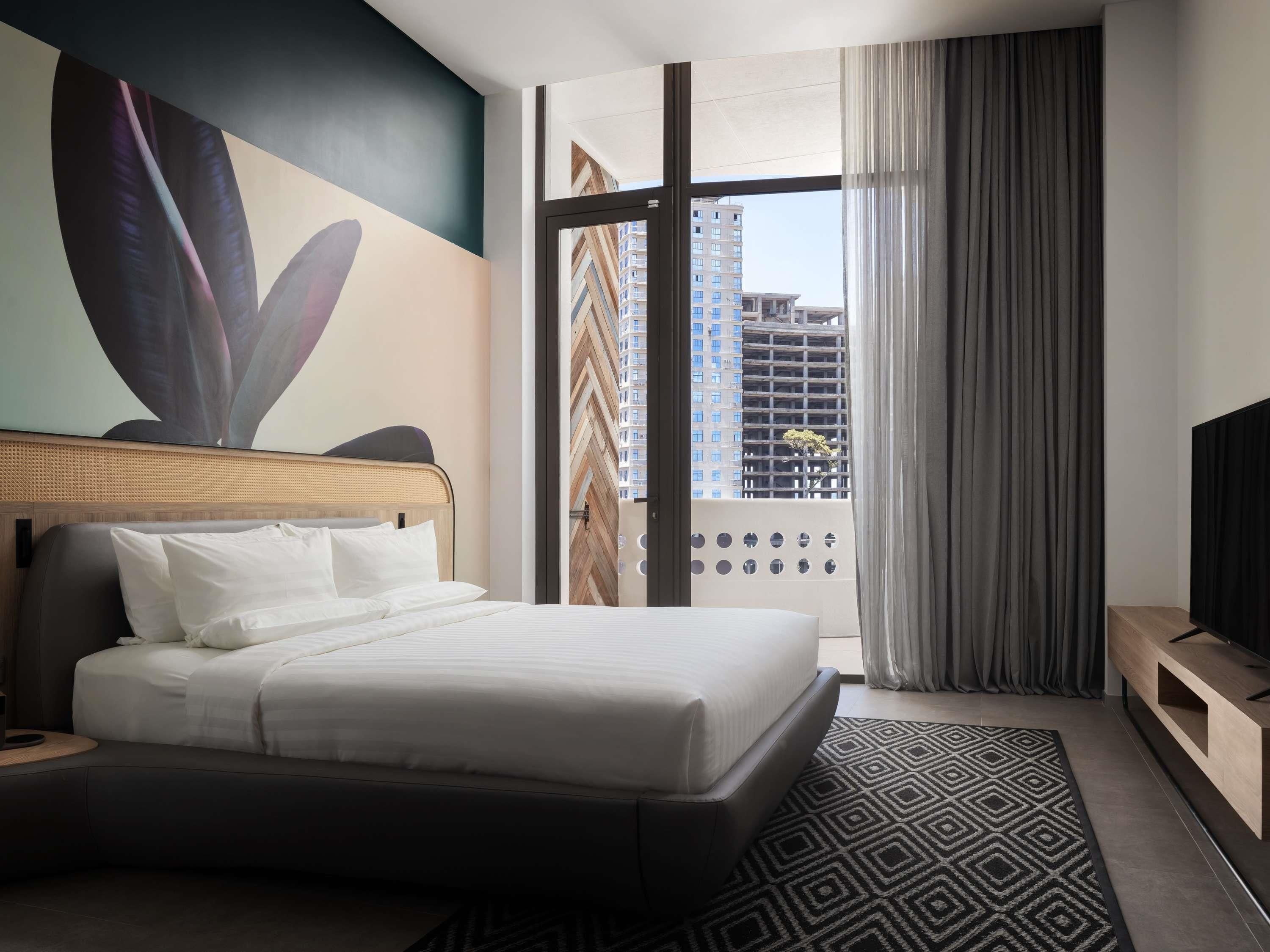 A hotel room with a bed, a large window, a view of buildings, a TV, and a wall with a geometric pattern.