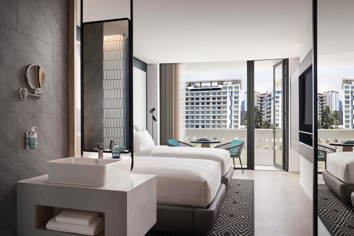 Une chambre d’hôtel moderne dotée d’un grand lit, d’une salle de bain séparée avec un meuble-lavabo, d’un balcon offrant une vue sur la ville et d’un grand miroir.
