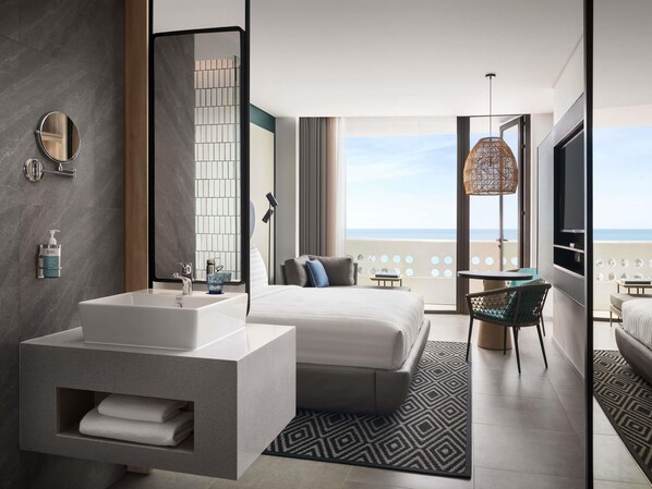 Habitación de hotel moderna con una cama grande, un baño con tocador, un balcón con vistas al mar y un televisor.
