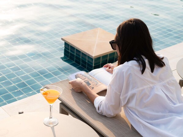 Una persona leyendo un libro junto a una piscina, con una bebida y una toalla.