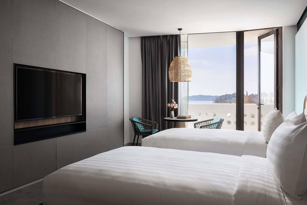 A hotel room with two beds, a TV, a view of the city, and a balcony with a table and chairs.