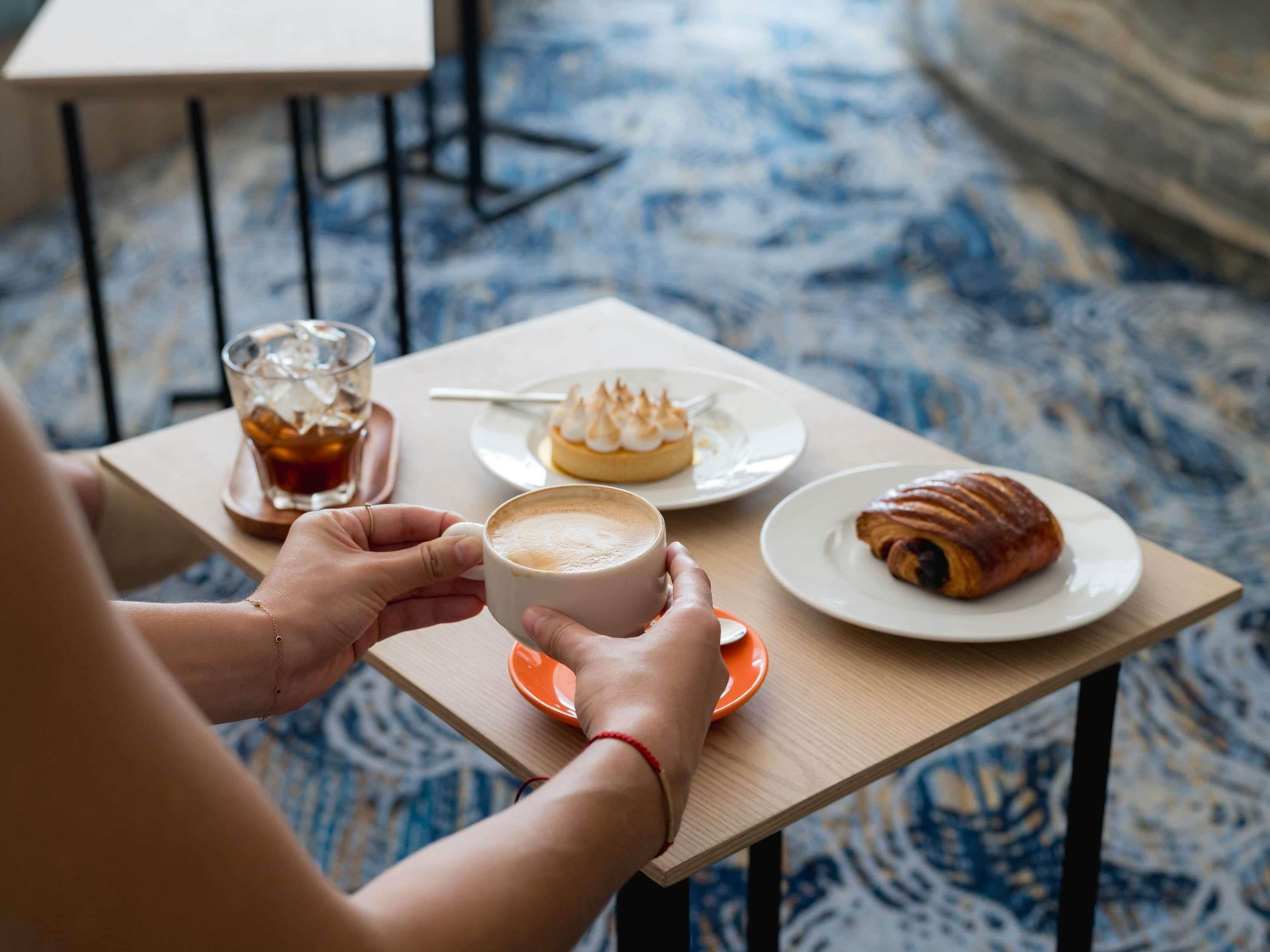 A person holding a cup of coffee at a table with a pastry and a glass of a dark beverage.