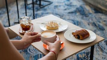 A person holding a cup of coffee at a table with a pastry and a glass of a dark beverage.