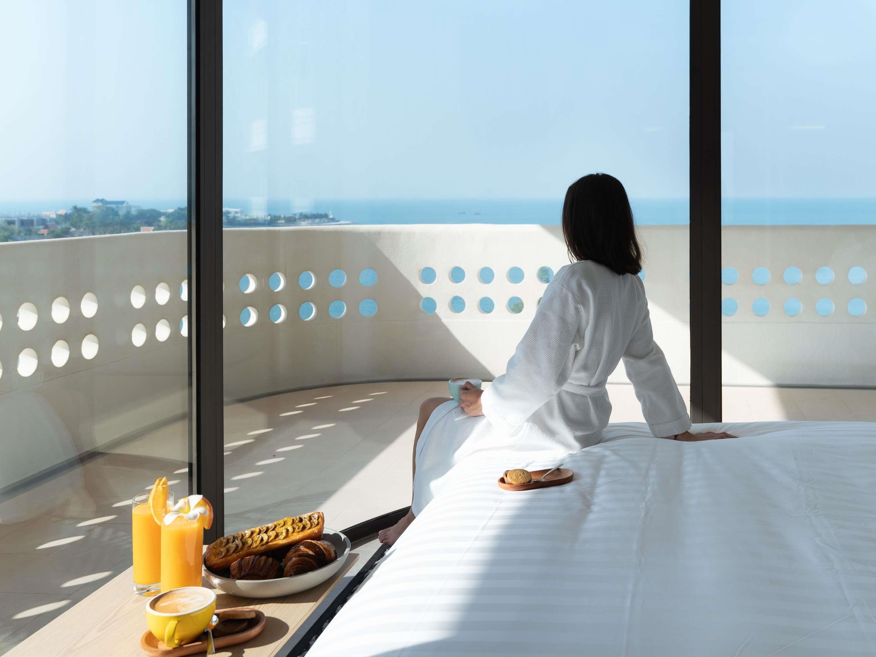 A woman sitting on a balcony with a bed, a breakfast tray, and a view of the sea.