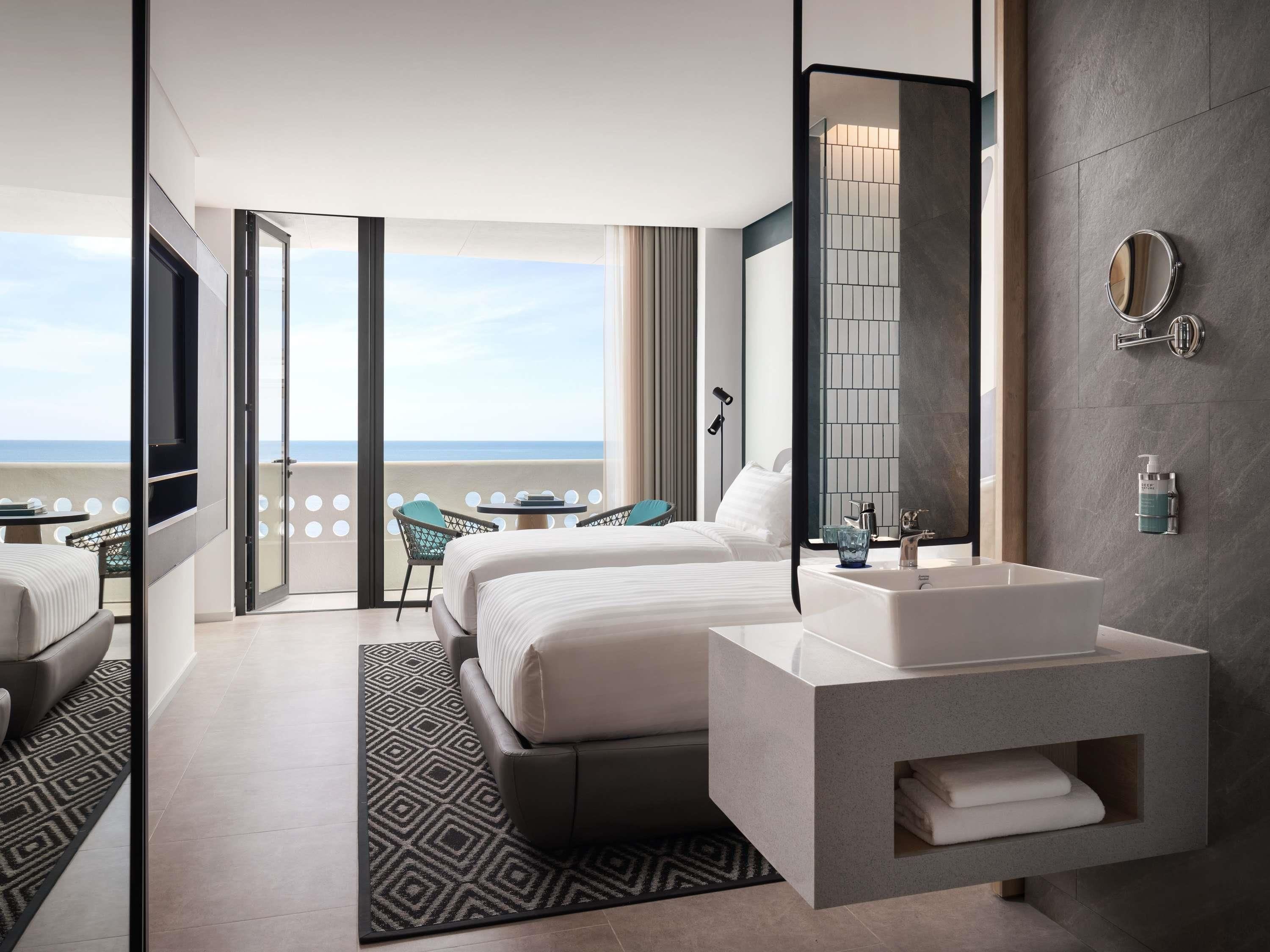 Ein modernes Hotelzimmer mit einem großen Bett, einem Badezimmer mit Waschbecken und Spiegel, einem Balkon mit Meerblick und einem gemusterten Teppich.