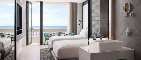 A modern hotel room with a large bed, a bathroom with a sink and mirror, a balcony with a view of the sea, and a patterned rug.