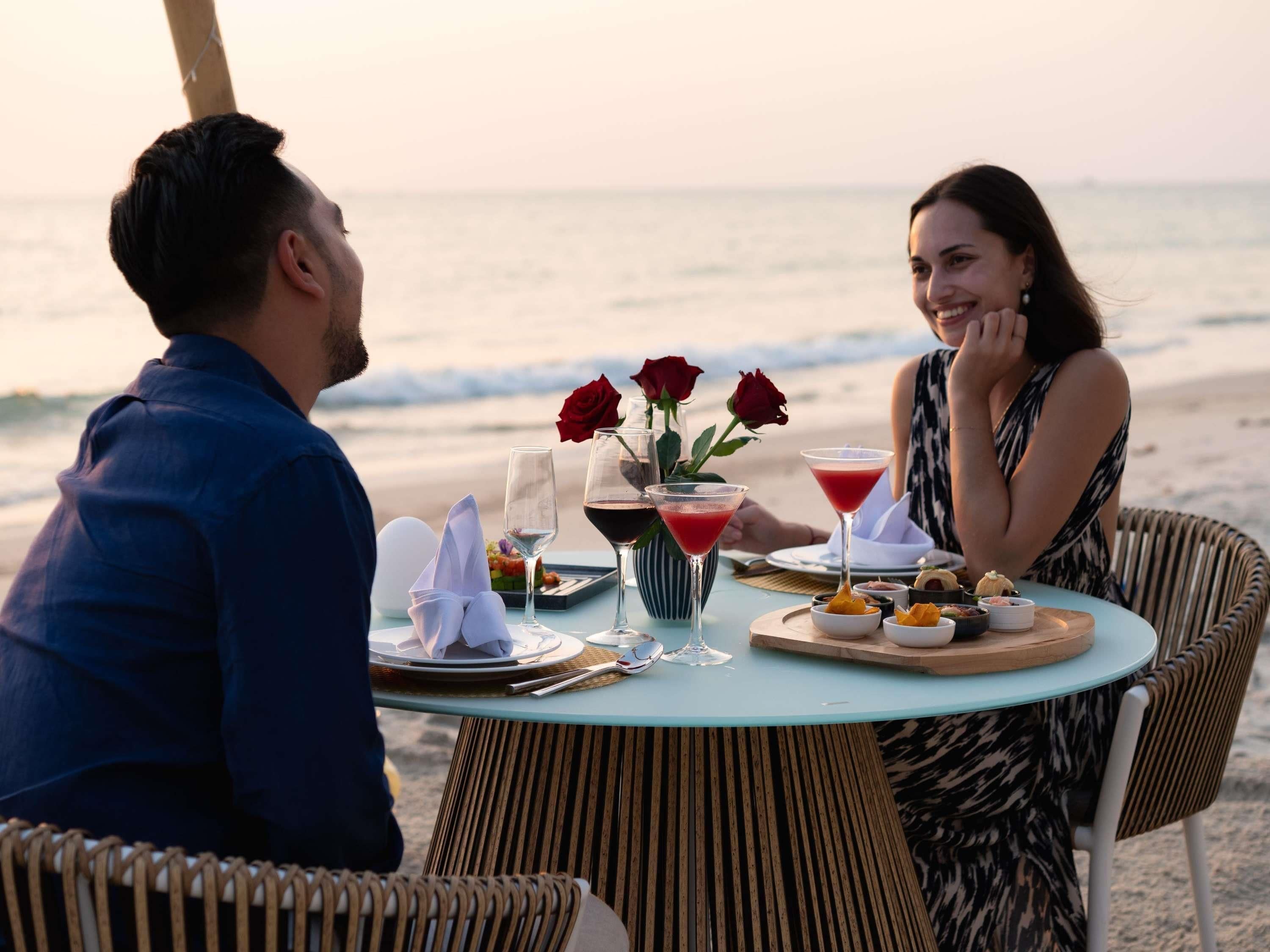 Een koppel dineert aan een tafel met uitzicht op de oceaan, compleet met een maaltijd, wijnglazen en een roos als middelpunt.
