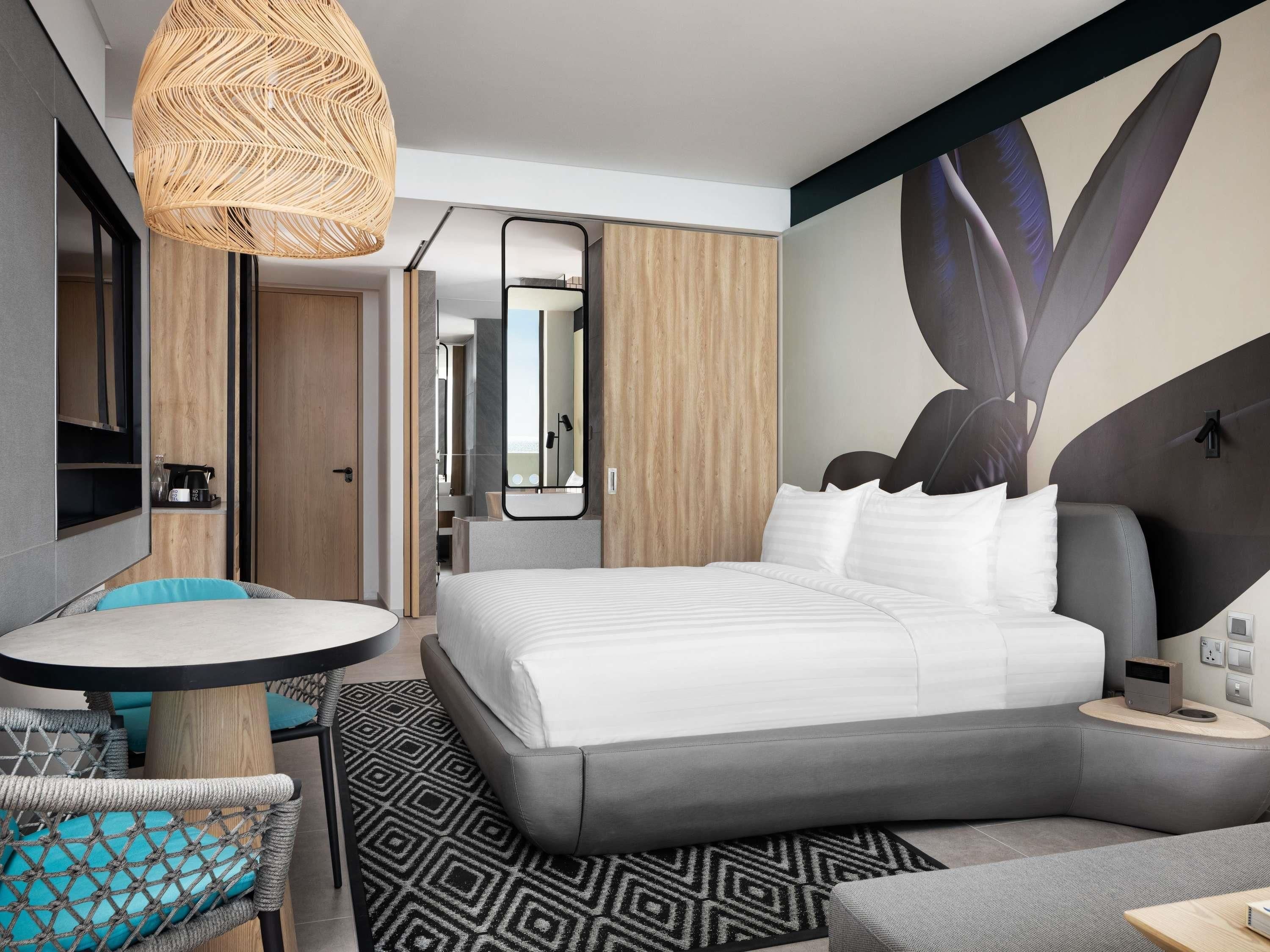 A modern hotel room with a bed, a round table, a chair, a large mirror, and a wall art piece.