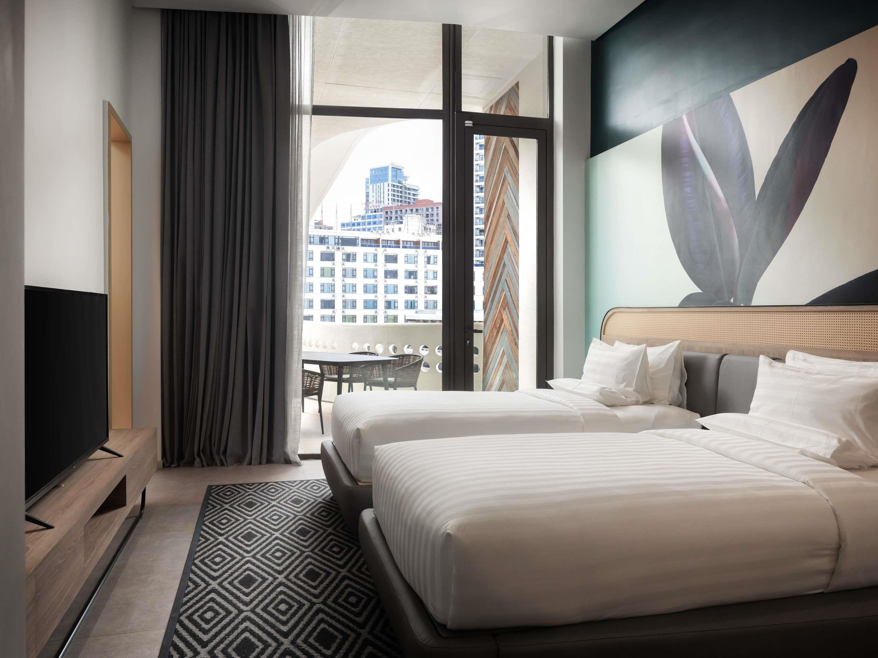 A hotel room with a bed, a TV, a dining table, and a view of a building.