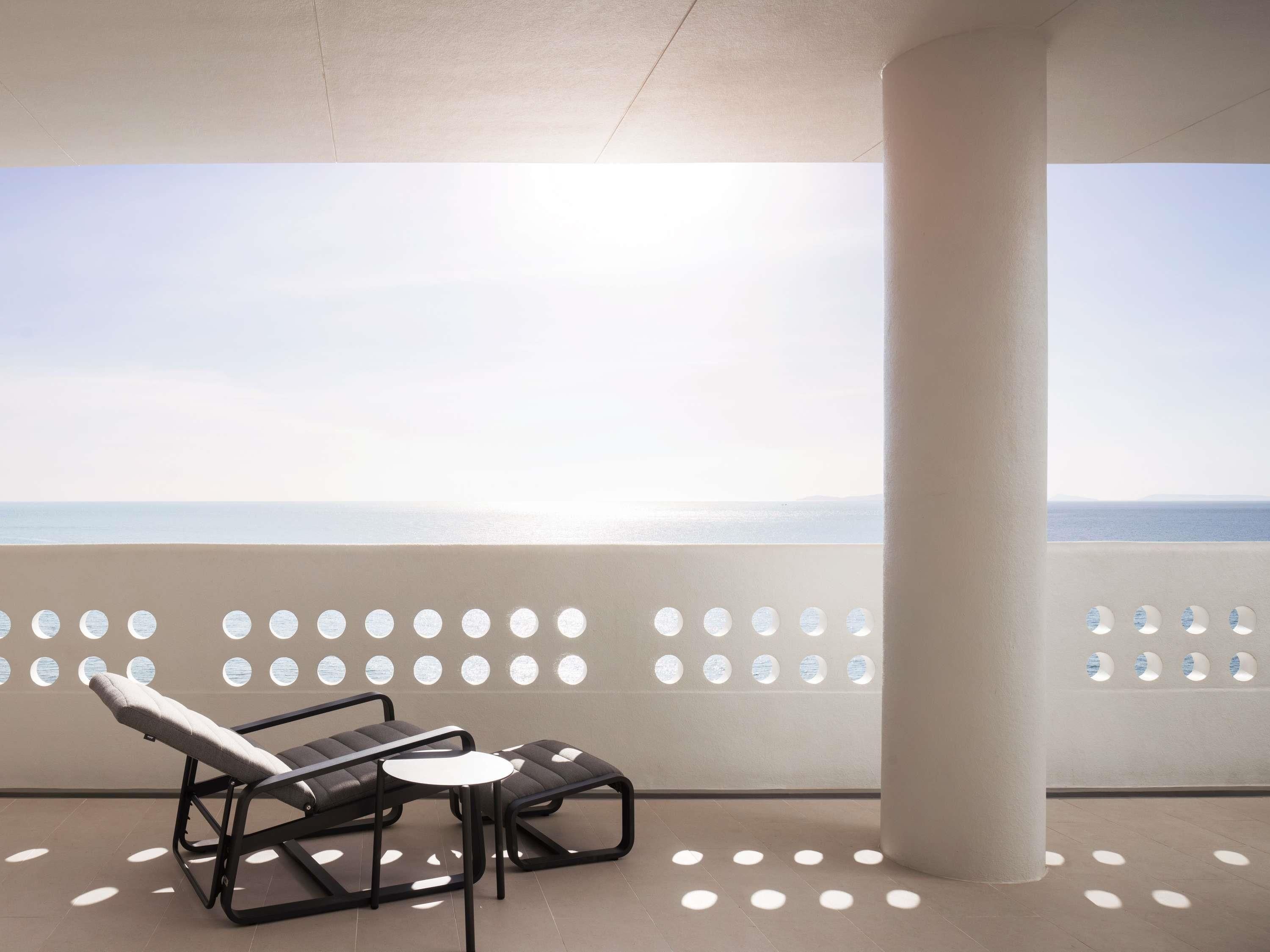 Un balcón con vistas al mar, una silla de descanso, una mesita y una columna blanca.