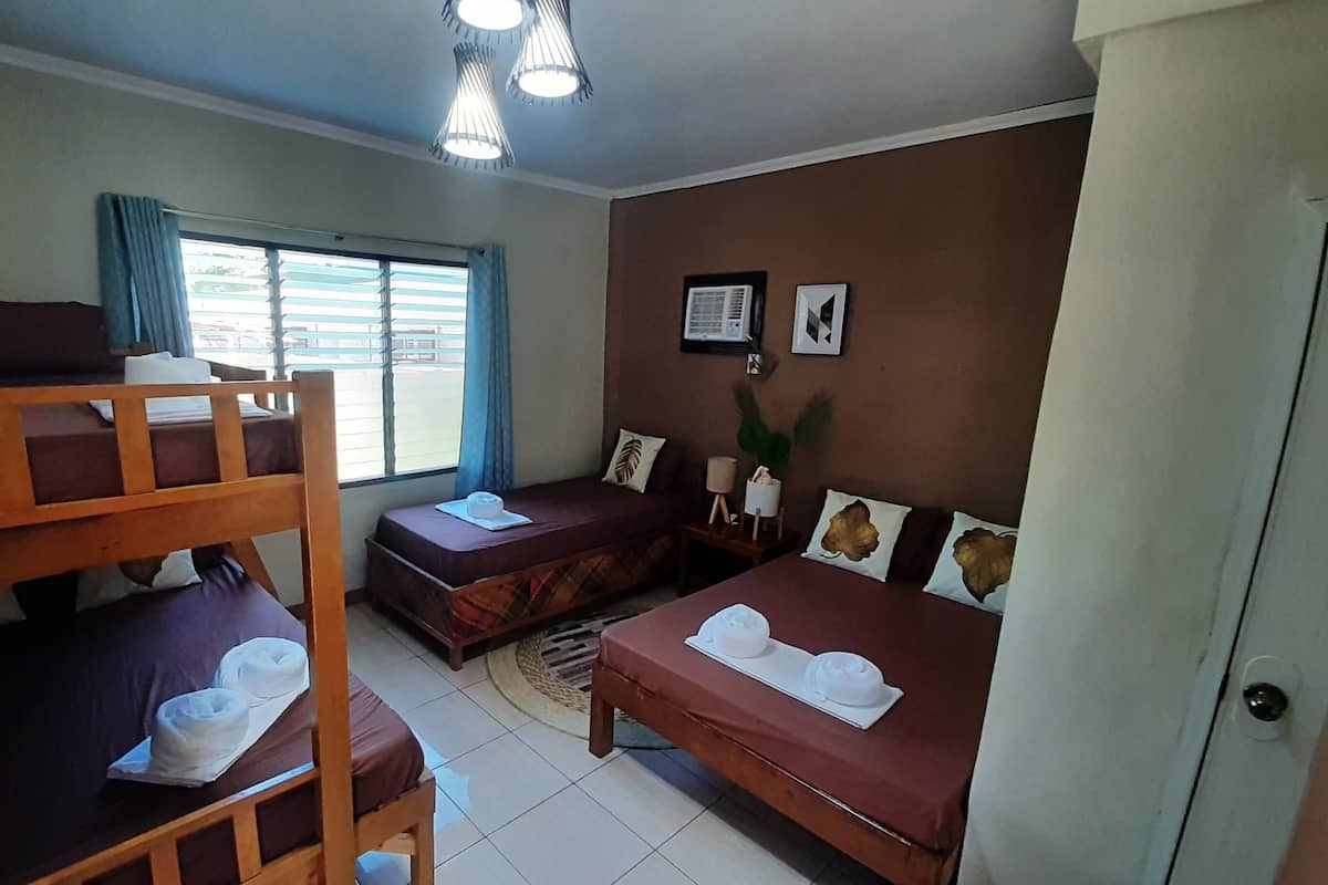 A dormitory room with bunk beds, a single bed, and a desk. There are towels on the beds, a window with curtains, and a wall-mounted air conditioner.