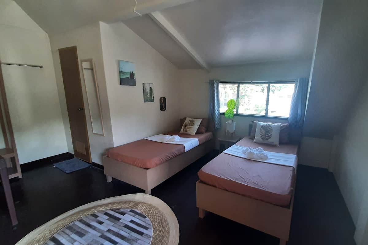 A small, cozy room with two single beds, a wooden chair, a small table, and a window with a view of greenery.