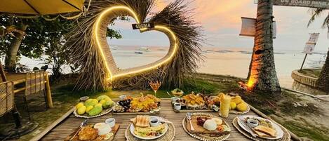 A wooden table set with various dishes and a heart-shaped light decoration at a beachside location.