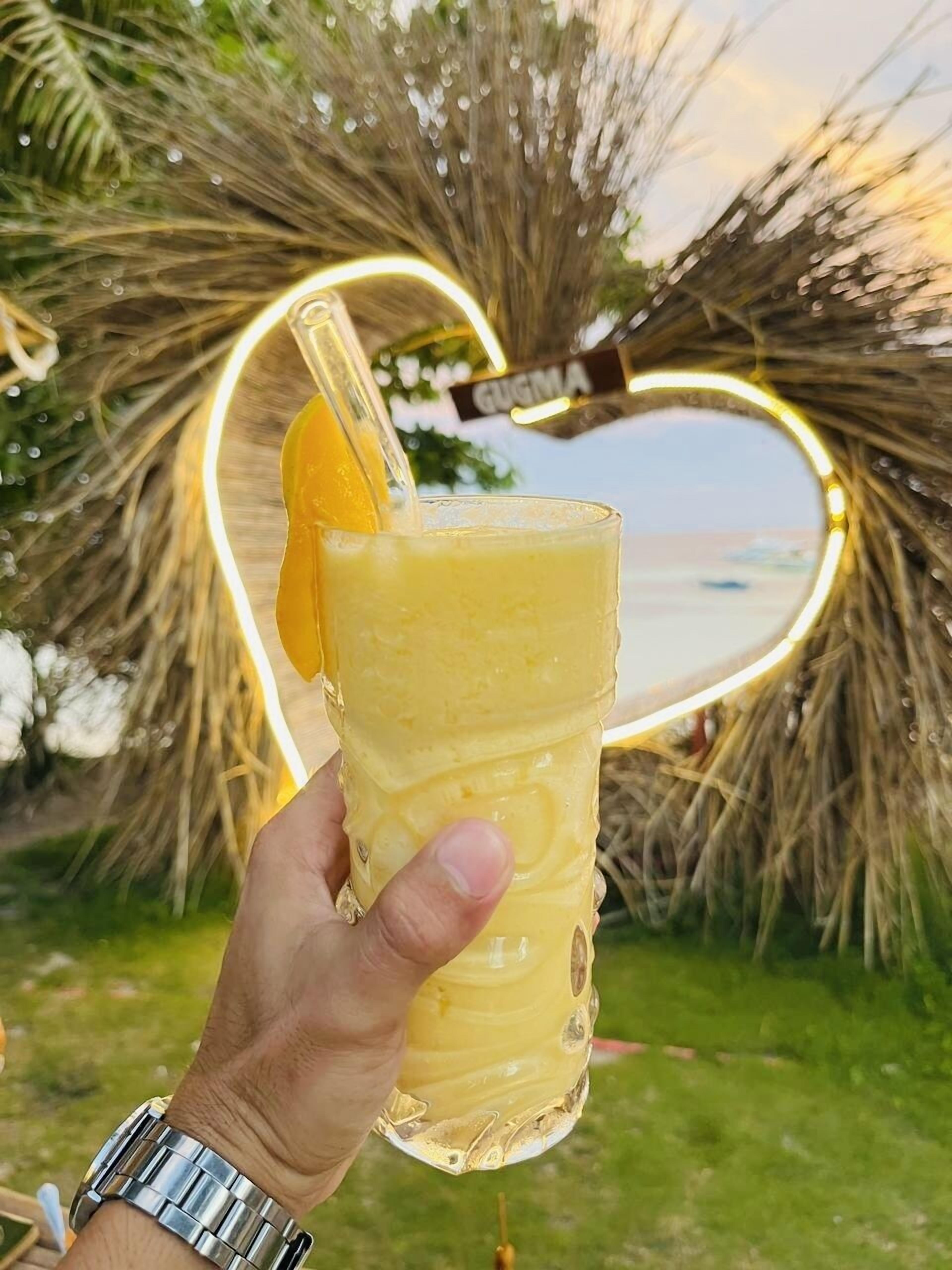 A hand holding a glass with a yellow beverage, palm leaves, and a heart-shaped light decoration in the background.