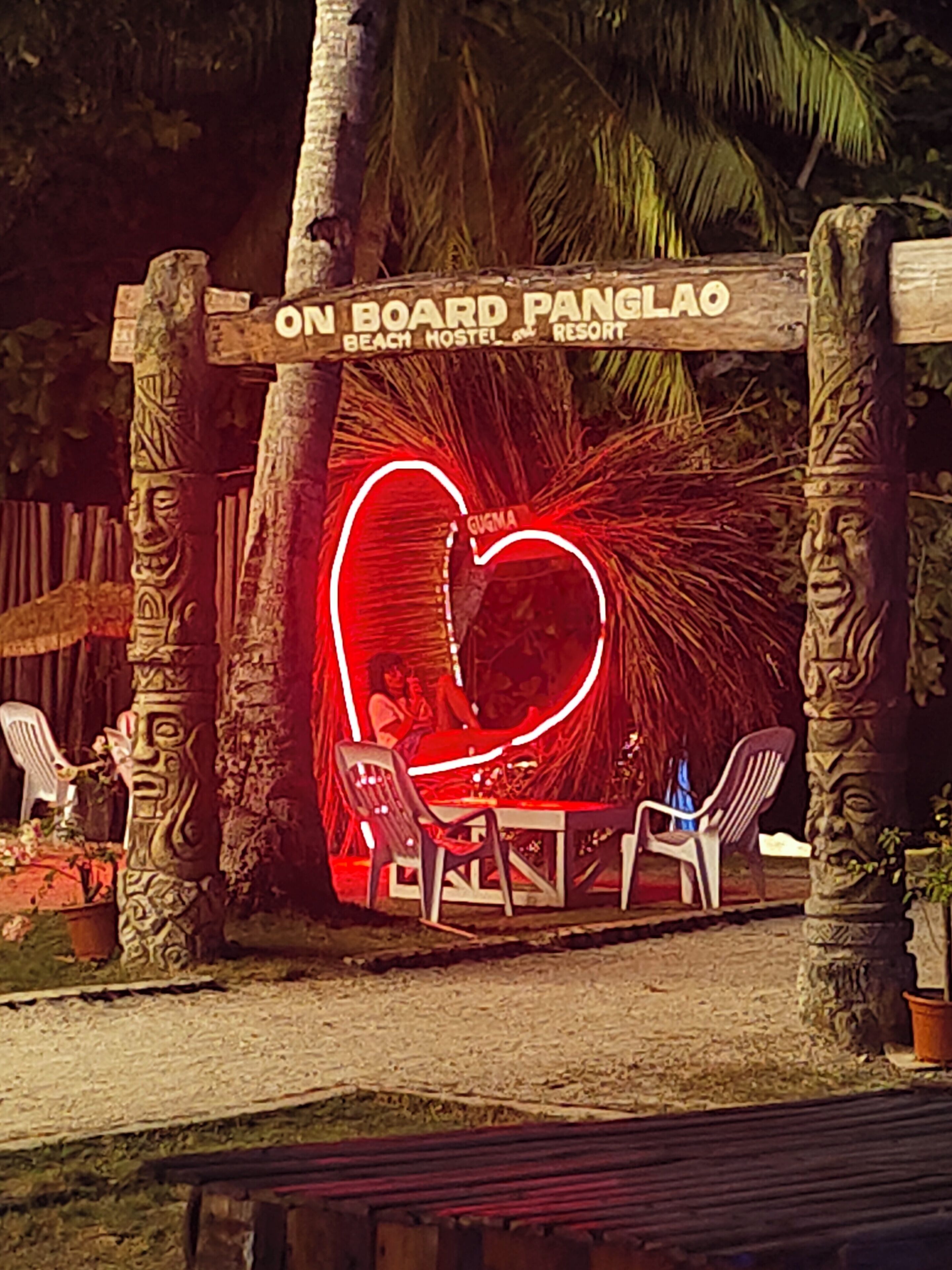 A beach hostel entrance with a neon heart sign, stone pillars, and plastic chairs.