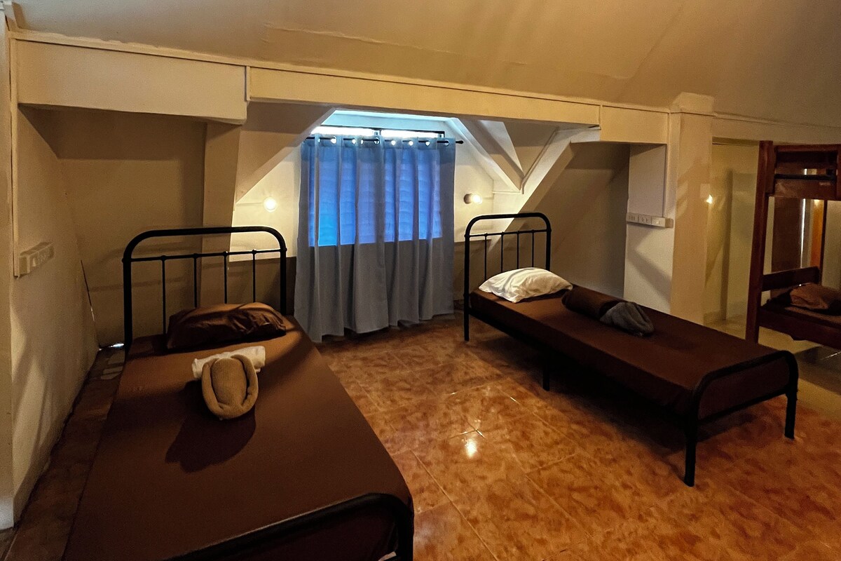 A dormitory room with two single beds, a window with curtains, and a sloped ceiling.