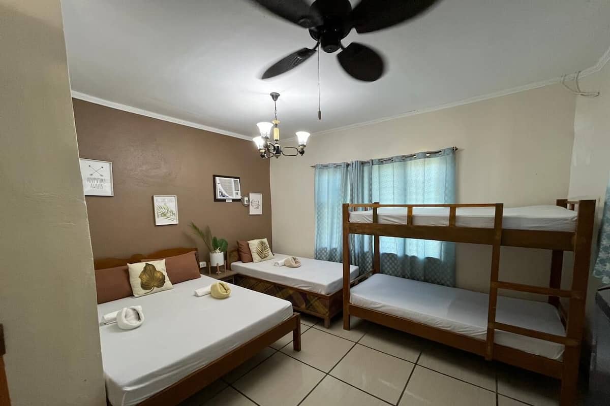 A hotel room with two single beds, a bunk bed, a ceiling fan, a chandelier, and a window with curtains.