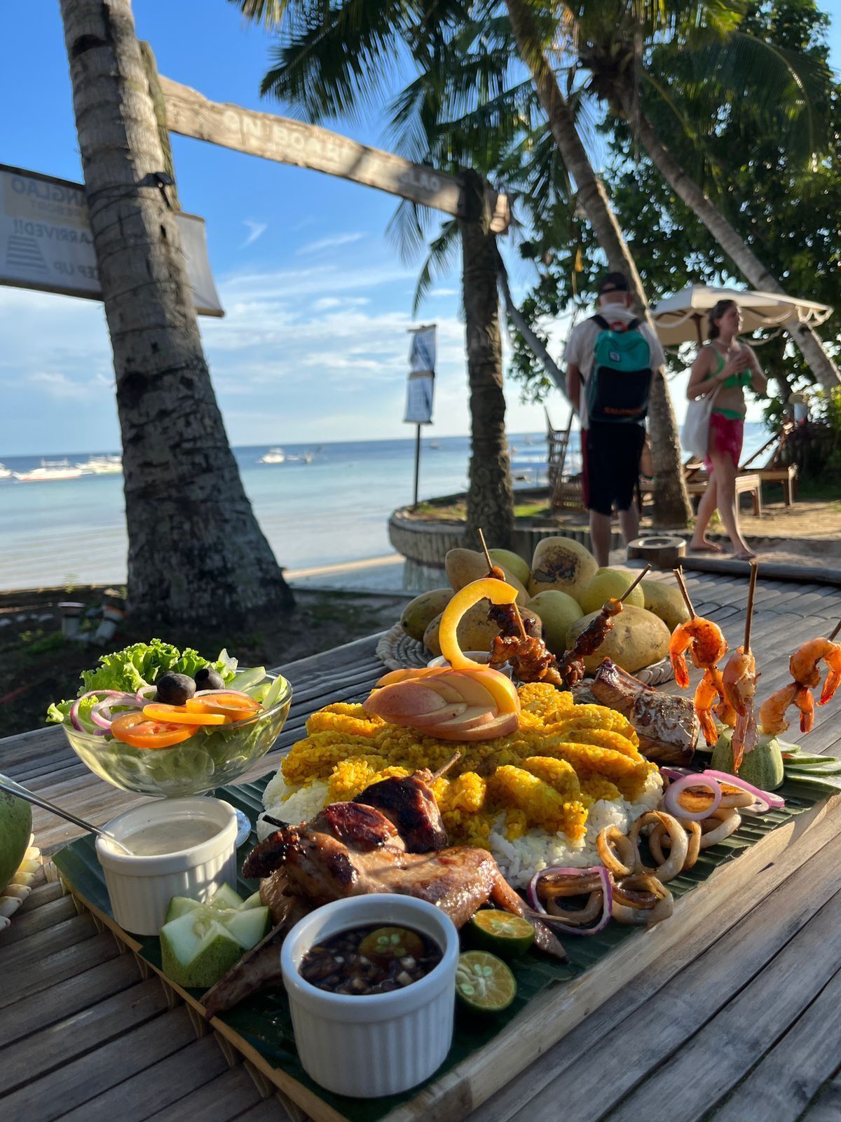 A wooden table with a variety of dishes including grilled meat, seafood, and rice, set on a wooden deck with a view of the ocean and palm trees.